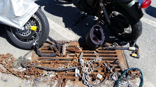 Two motorcycles locked to a rusty storm drain by a tangled assortment of chains 