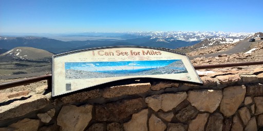 A signboard at the summit of Mount Evans against a backdrop of mountains and blue sky helps visitors identify noteworthy peaks   